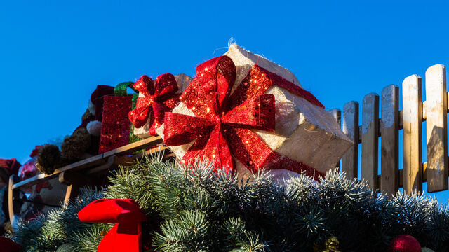 Geschenk_mit_Schlitten Ein auf Tannenzweigen präsentiertes goldenes Geschenk mit roter Schleife vor blauem Himmel im Winter